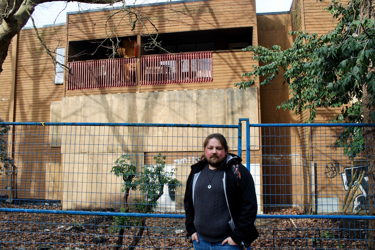 A light-skinned man with shoulder-length brown hair is wearing a blue sweater and looking directly at the camera. He has his hands in his pockets. Behind him is the boarded-up, fire-damaged Mount Pleasant building. 