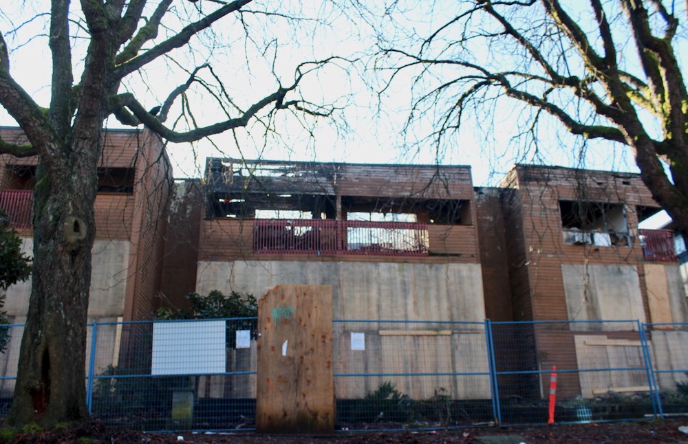 Blue fences and orange traffic cones wrap around a fire-damaged wood-panelled apartment building that is visibly charred and fire-damaged. 