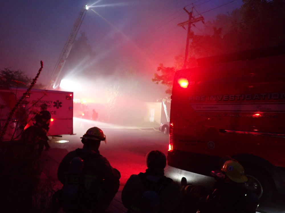 Nighttime. Silhouettes of firefighters are visible via the lights from a fire vehicle and an EMS vehicle. 