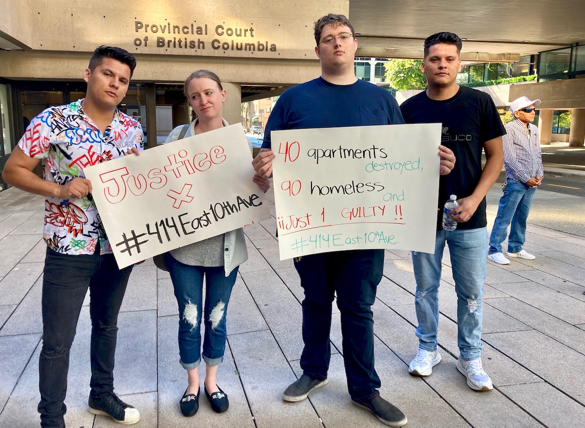 Four people stand next to each other, holding two signs. One says, &lsquo;Justice X #414East10thAve.&rsquo; The other says, &rsquo;40 apartments destroyed, 90 homeless, just 1 guilty!&rsquo;