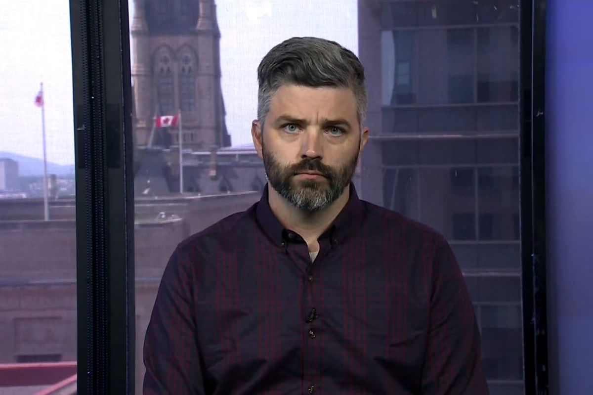 An early-middle-aged man with light skin and salt and pepper hair speaks in a room with the Parliament Buildings and Canadian flag in the background out a window.