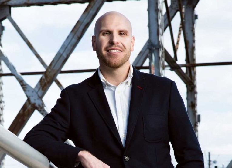 A headshot of a man with a bald head wearing a white button-down shirt and black blazer. The man is looking at the camera and smiling.