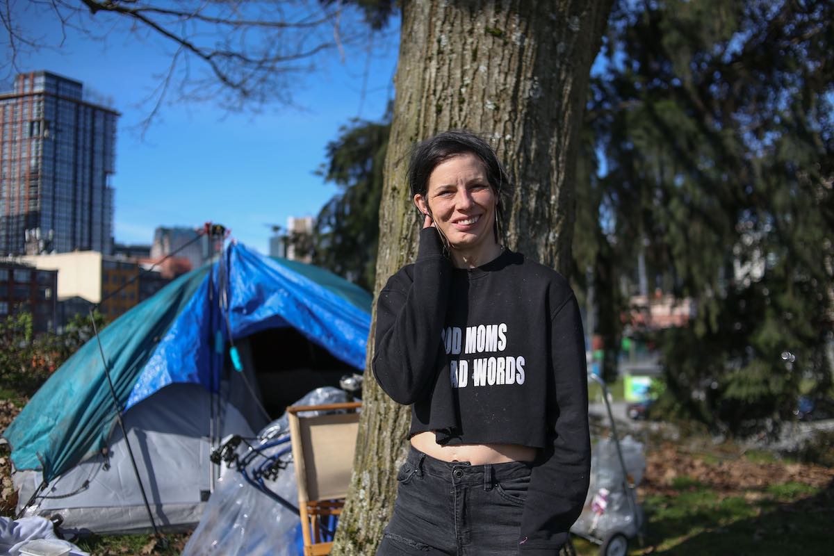 A young woman with dark hair stands in front of a tent wearing dark jeans and a black T-shirt.