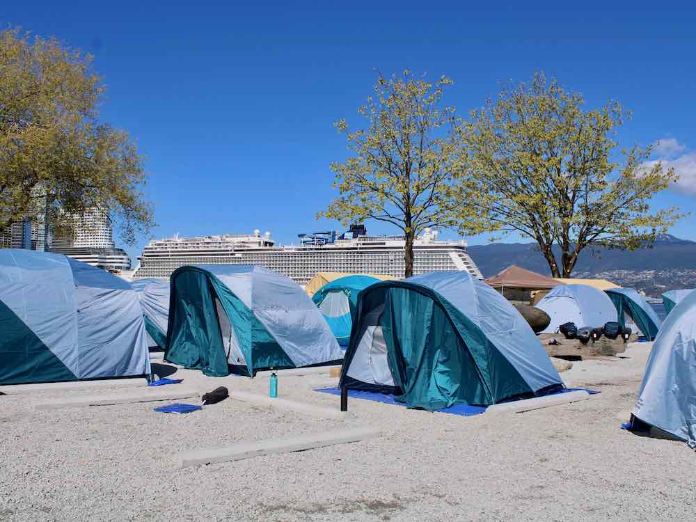 Blue tents are lined up on a gravel pad with concrete parking lot barriers separating them. In the background is a cruise ship.