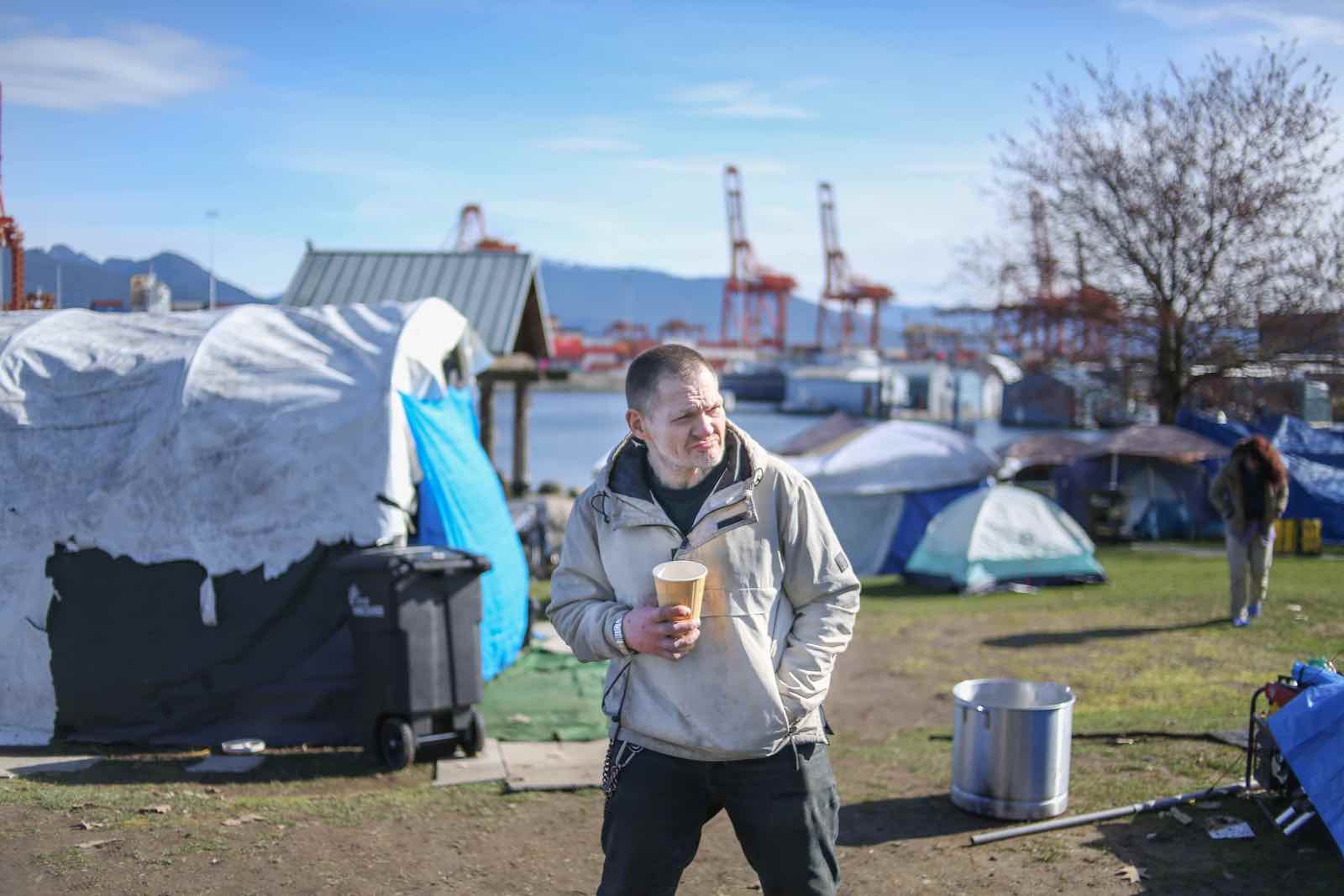 A man with short hair stands in a camp area, holding a coffee cup.