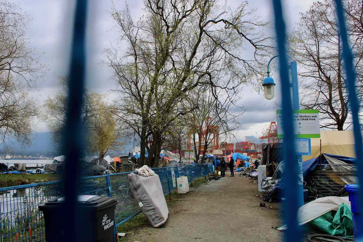 City workers stand in the distance. A garbage can and mattress and tents are in the foreground.