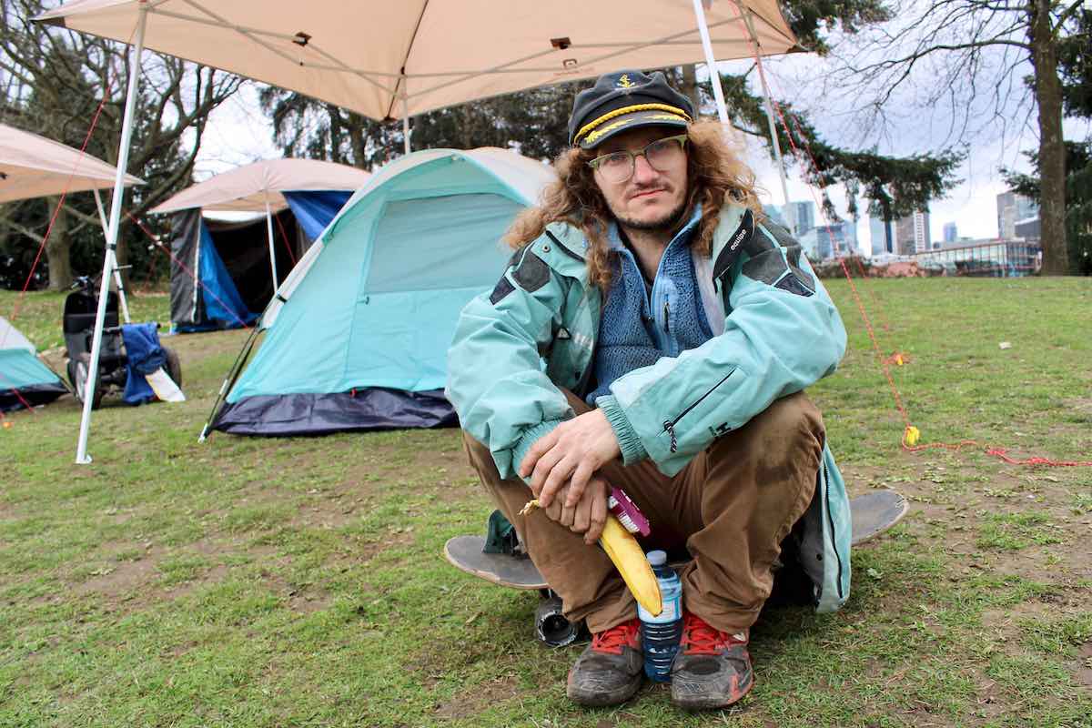 A person with long brown hair sits on a skateboard in front of a tent, wearing a light blue windbreaker and a captain’s hat and holding a banana.