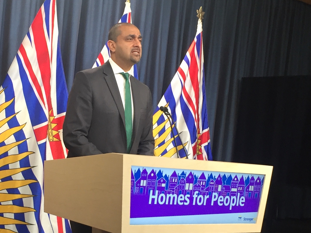 A 44-year-old man with medium-toned skin stands at a podium with a sign saying 'Homes for People.' He wears a grey suit, white shirt and green tie.