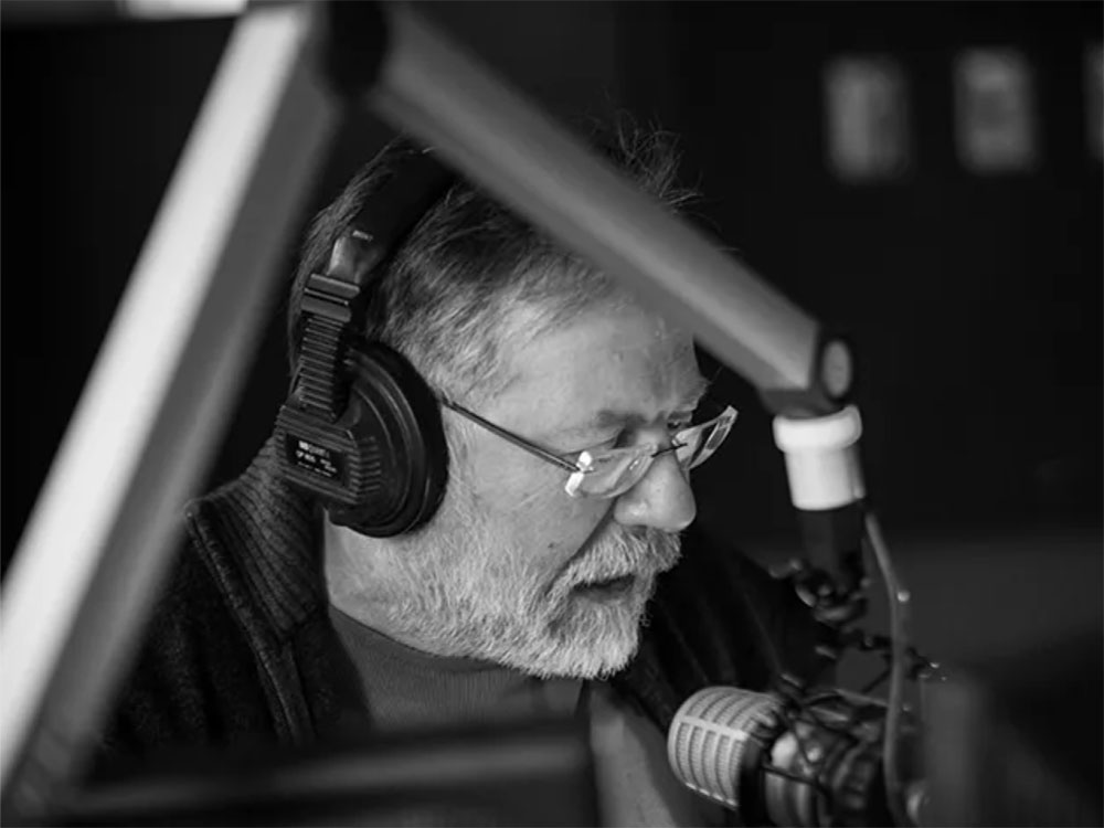 A black and white photo of a man with grey hair and beard and frameless glasses, wearing headphones and speaking into a microphone in a recording studio.