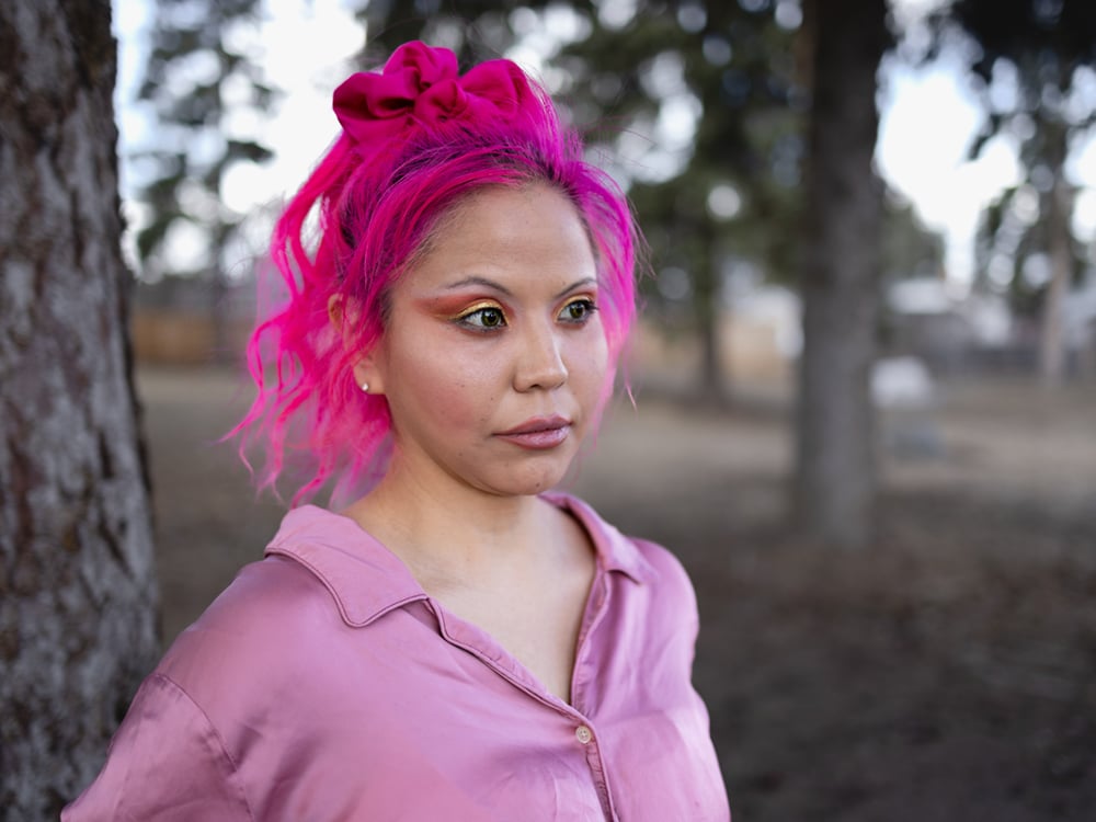 A woman with pink hair and a pink button-down shirt stands with trees behind her, looking past the camera.