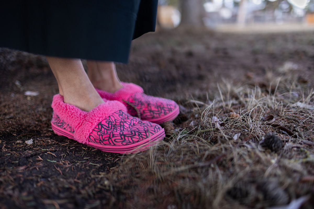 A close-up image of a pair of feet wearing pink and black fuzzy slippers and standing on ground covered with earth and pine needles.