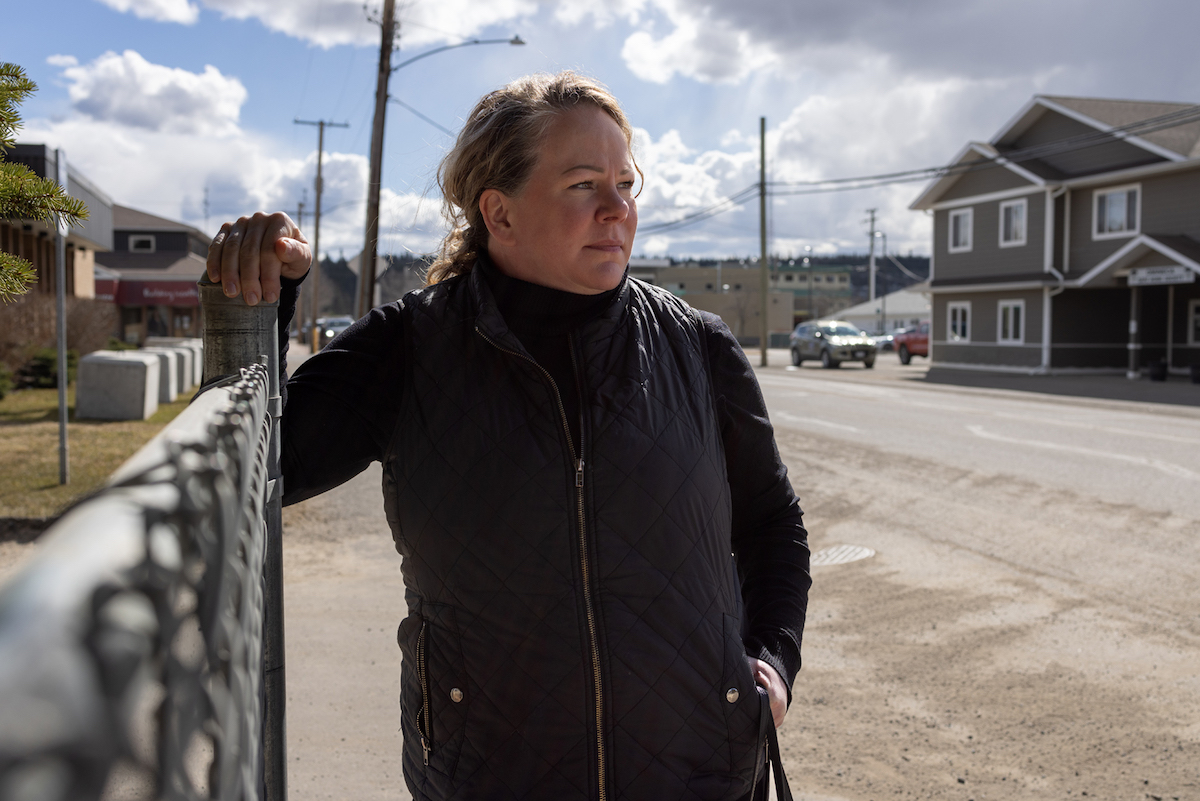 A woman with blond hair tied back in a ponytail, and wearing a black shirt and vest, stands next to the street with one hand resting on a chain-link fence. She is looking past the camera.