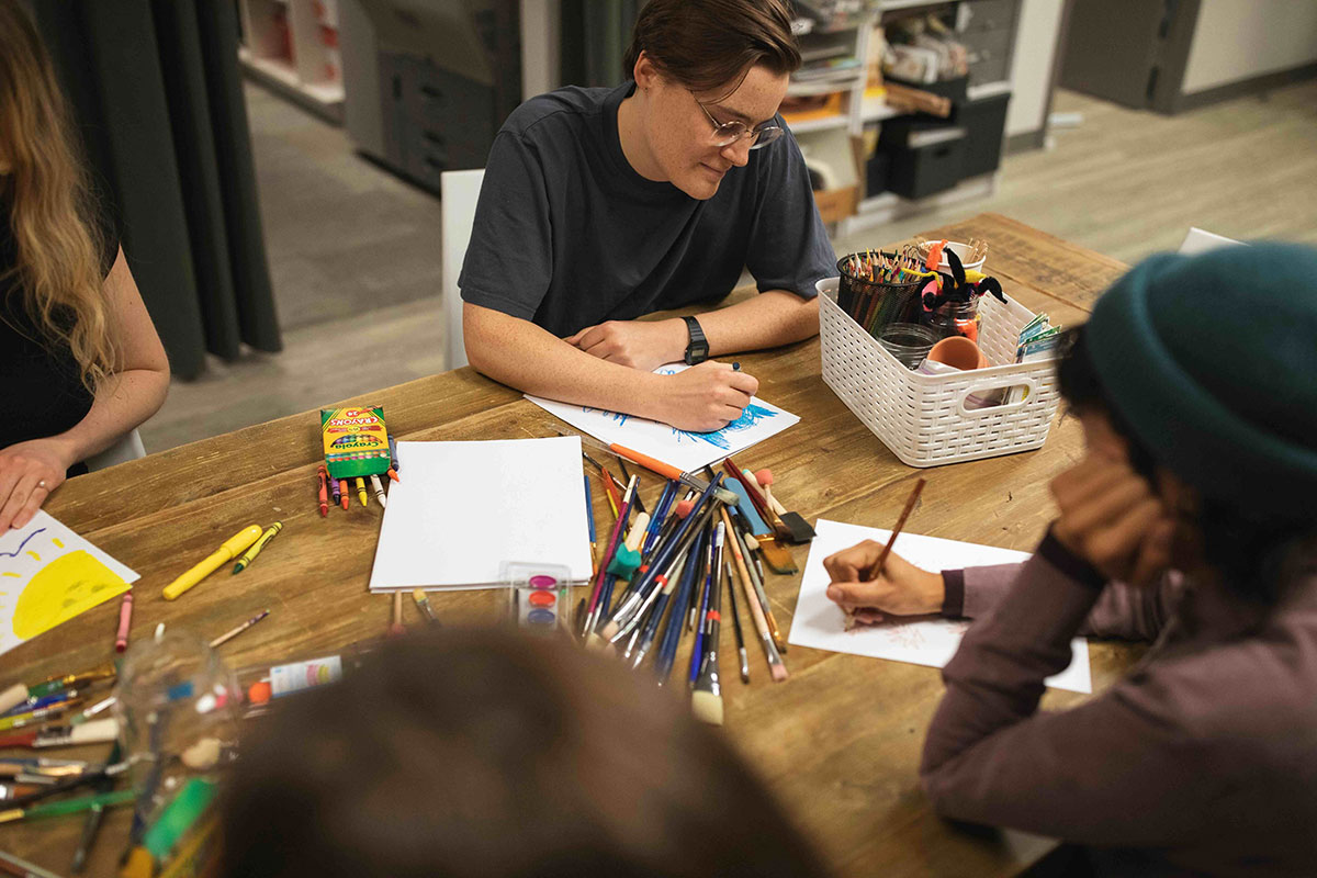 People are seated around a wooden table equipped with art supplies. They are writing and drawing on white paper.