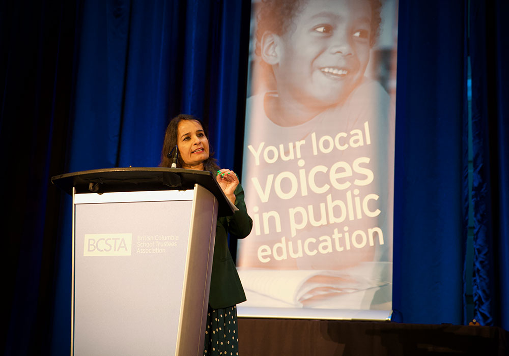 A petite woman with medium-light skin tone and straight, dark brown hair stands at a podium in front of a vertical banner that reads, ‘Your Local Voices in Public Education.’