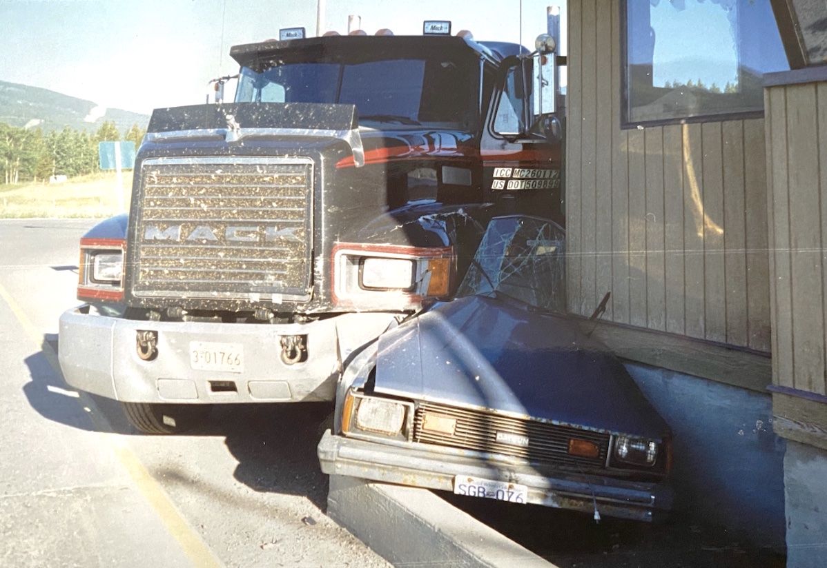 A black Mack semi has crushed a blue car against a building.