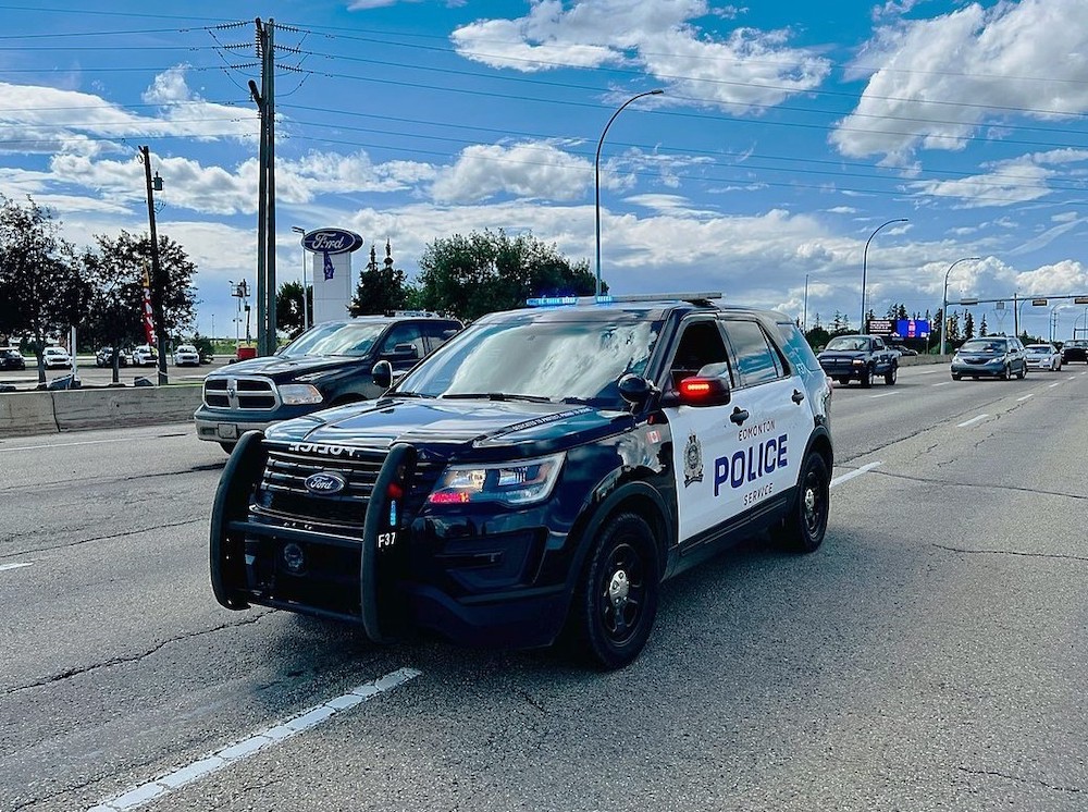 A black and white Edmonton Police Service car drives among other cars on a freeway under a cloud-dotted blue sky.