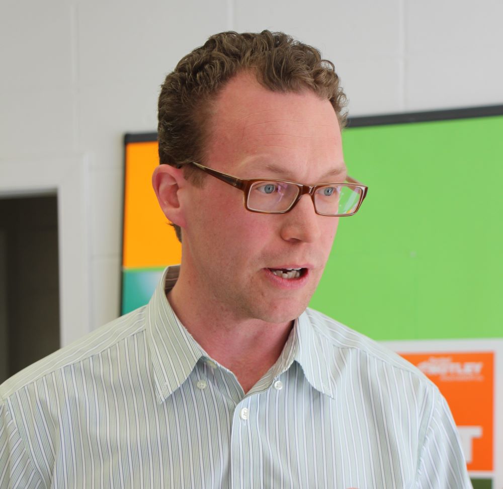 A light-skinned man with short brown hair, blue eyes and brown-framed glasses, wearing a striped light-coloured collared shirt, looks to the right of the frame, appearing to be in mid-speech.