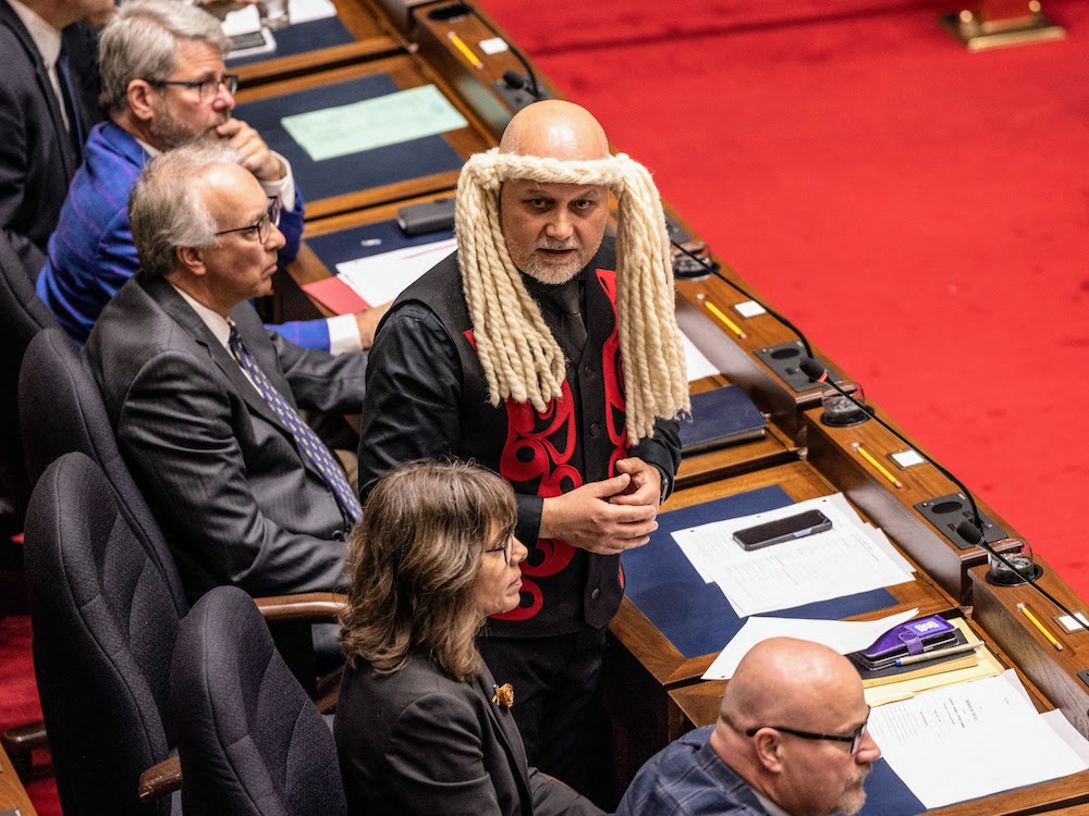 An elevated view shows a bald man with a grey goatee standing alongside others sitting at linked wooden desks on a red carpet. He wears a straw-coloured headband with long braids on each side and a black vest with red figures.