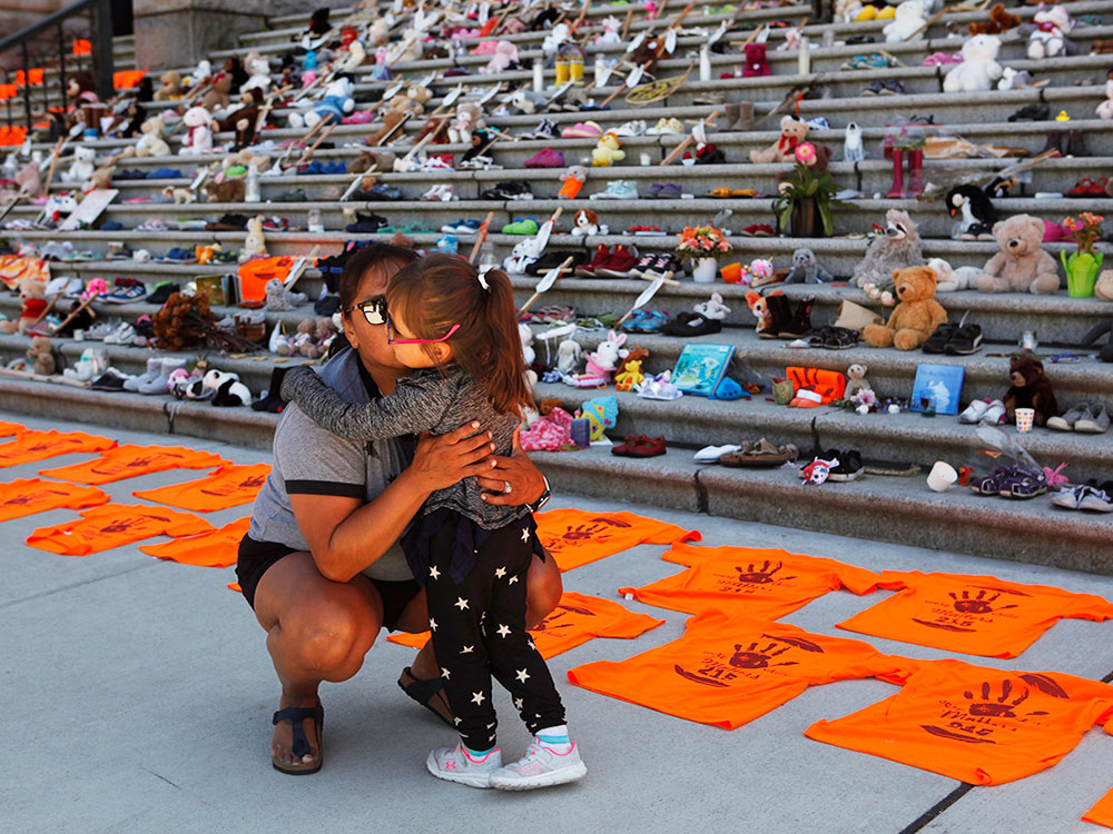 An Indigenous woman squats down, hugging a child. Behind the are orange shirts and steps covered with small shows and teddy bears.