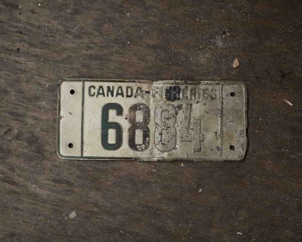 A warped licence plate with the words 'Canada Fisheries' across the top is laid on a brown tabletop.