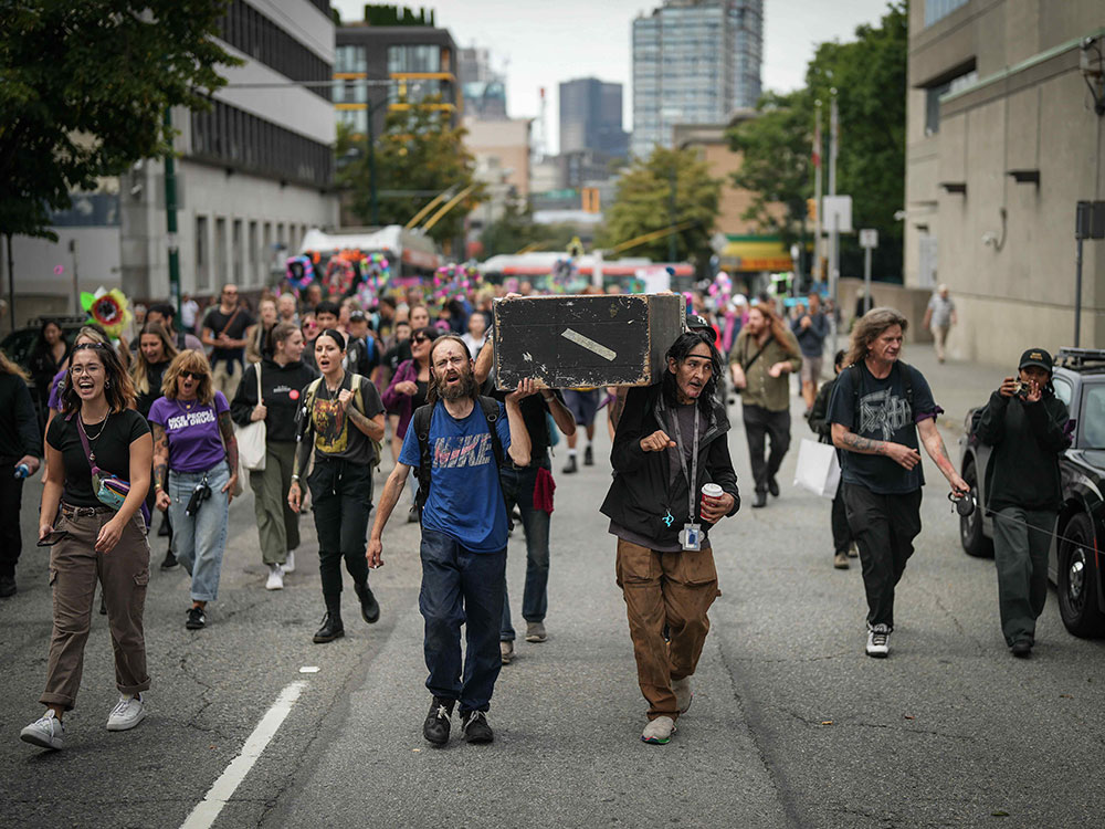 People march in a street. In the front, two men hold a battered black coffin on their shoulders.