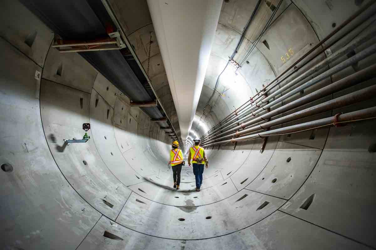 Two workers in fluorescent yellow construction hats and white helmets walk with their backs to the camera in a large grey concrete tunnel. A large white pipe spans across the top of the tunnel and several thinner pipes run to the workers&rsquo; right.