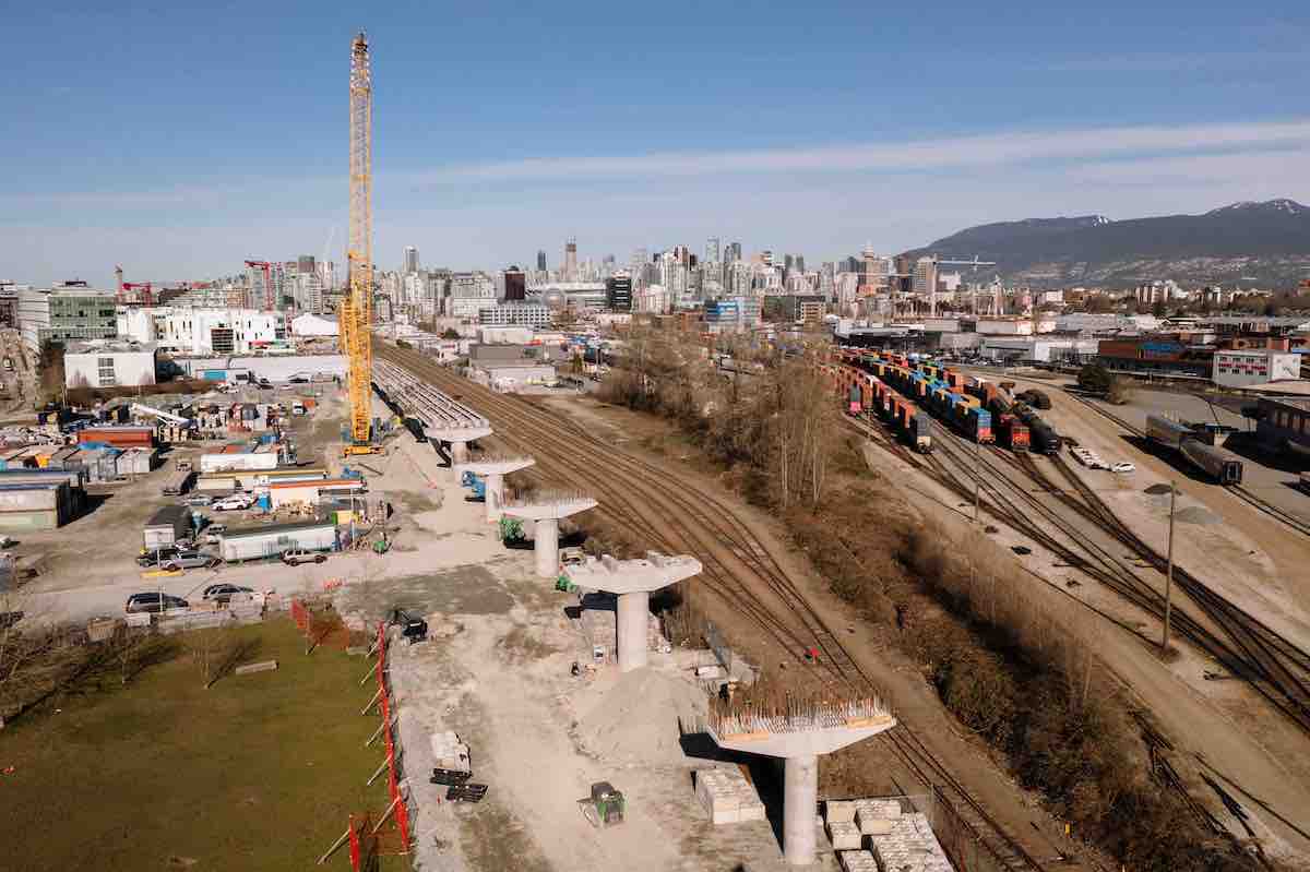 An elevated view of a row of several raised concrete platforms for the future Broadway Subway line that run alongside train tracks to their right. A yellow construction crane is to the left of the platforms. It&rsquo;s a bright winter day.