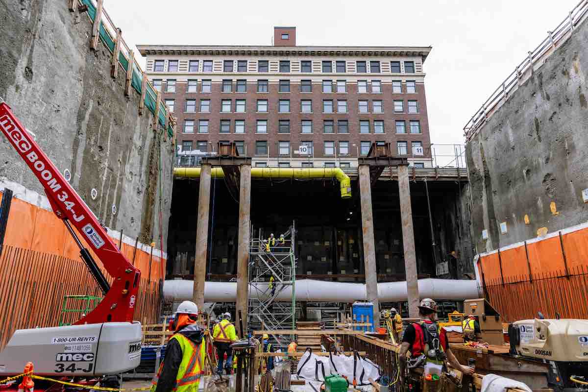 Construction crews work below ground in high-visibility vests among tall vertical pipes and equipment. Just above ground is a tall heritage brick building with rows of small rectangular windows.