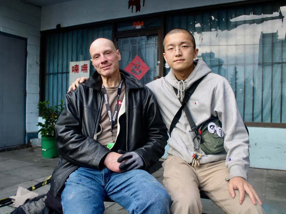 Two people are sitting outside, facing the camera. On the left, a light-skinned man with no hair is wearing a black leather jacket. On the right, a person with medium-toned skin and a buzz cut is wearing a grey hoodie and glasses.