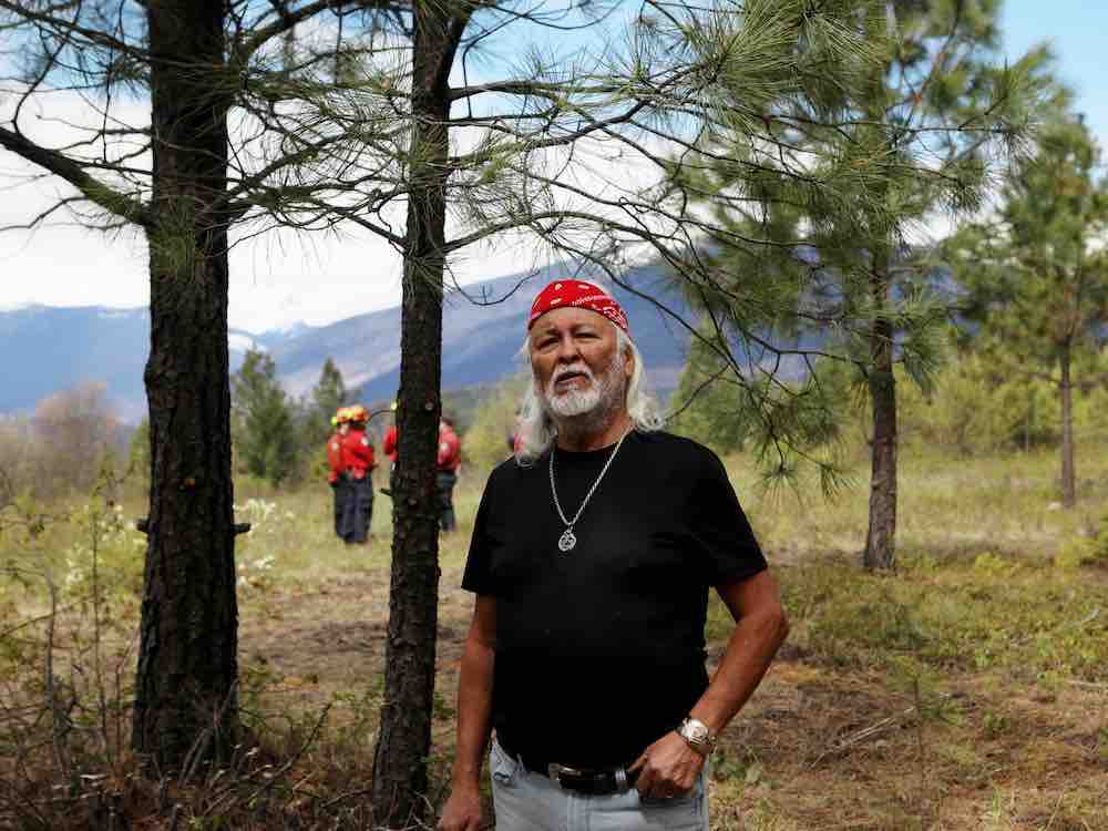 Elder Rick Campbell is an Indigenous man with medium skin, shoulder-length white hair and a beard. He wears a black T-shirt, light blue jeans and a red bandana around his head. He’s standing in a forest and behind him is a wildfire service crew in red uniforms.