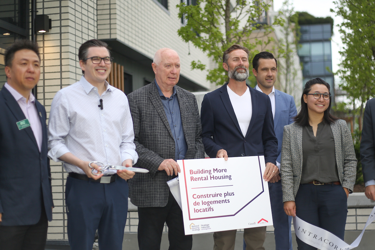 A group of dignitaries stand in front of a new building holding up a sign that says 'Building more rental housing.' One of the men holds a pair of giant scissors.