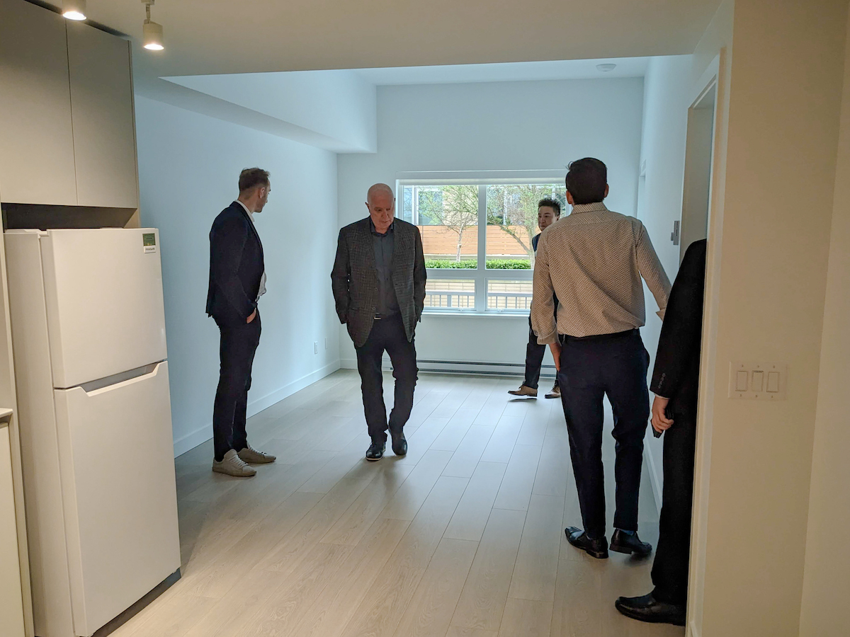Men in suits tour a new apartment unit. There is a view of landscaping outside the window.