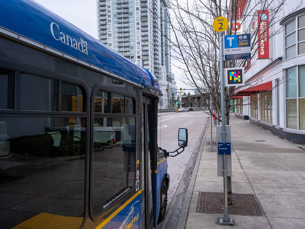 At a bus stop, there is a scannable sign of randomly arranged square pixels, coloured yellow, pink, blue and black, and a white-coloured border. A blue TransLink shuttle waits at the stop. 