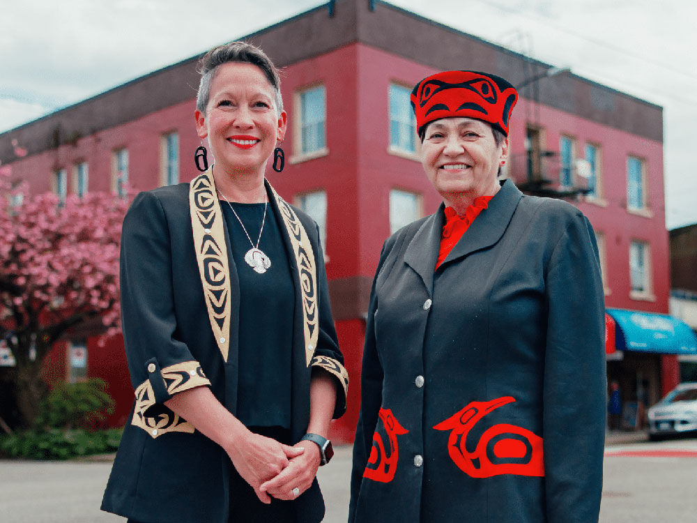 Two women, Melanie Mark and Joan Phillip, stand in front of a burgundy building. They are wearing black suits with Indigenous artistic detail.