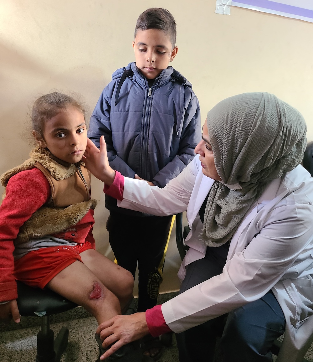 Fozia Alvi, right, wears a white lab coat and sage green head covering. She touches the cheek of a school-aged girl seated on a rolling office chair. She is wearing red and her leg is wounded. Behind her an older boy stands looking at her in a blue winter jacket.