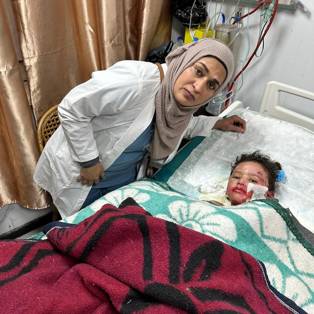 Fozia Alvi wears a white lab coat and a dusty rose head covering. She leans over a child wrapped in blankets in a hospital bed. The child has wounds on their face.