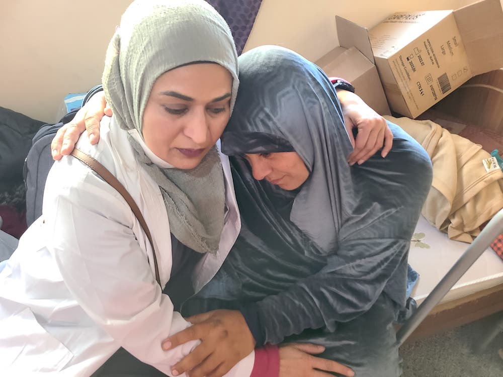 Fozia Alvi, left, wears a white lab coat and a sage green head covering. She embraces a woman on her right wearing a blue head covering and matching outfit. They are seated indoors on a hospital bed. Behind them are blankets and cardboard boxes.