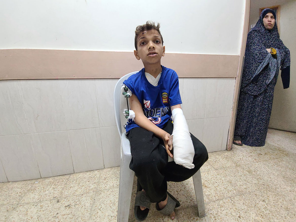A school-aged boy with short brown hair, medium skin and a blue sports T-shirt sits on a white plastic chair in a corridor with white walls and fluorescent lighting. One arm is in a cast and the other is held straight with a splint.