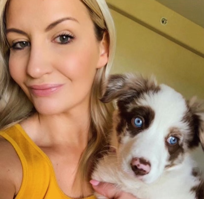 A young blond woman with light skin tone and green eyes looks at the camera, holding a fluffy Australian shepherd puppy.