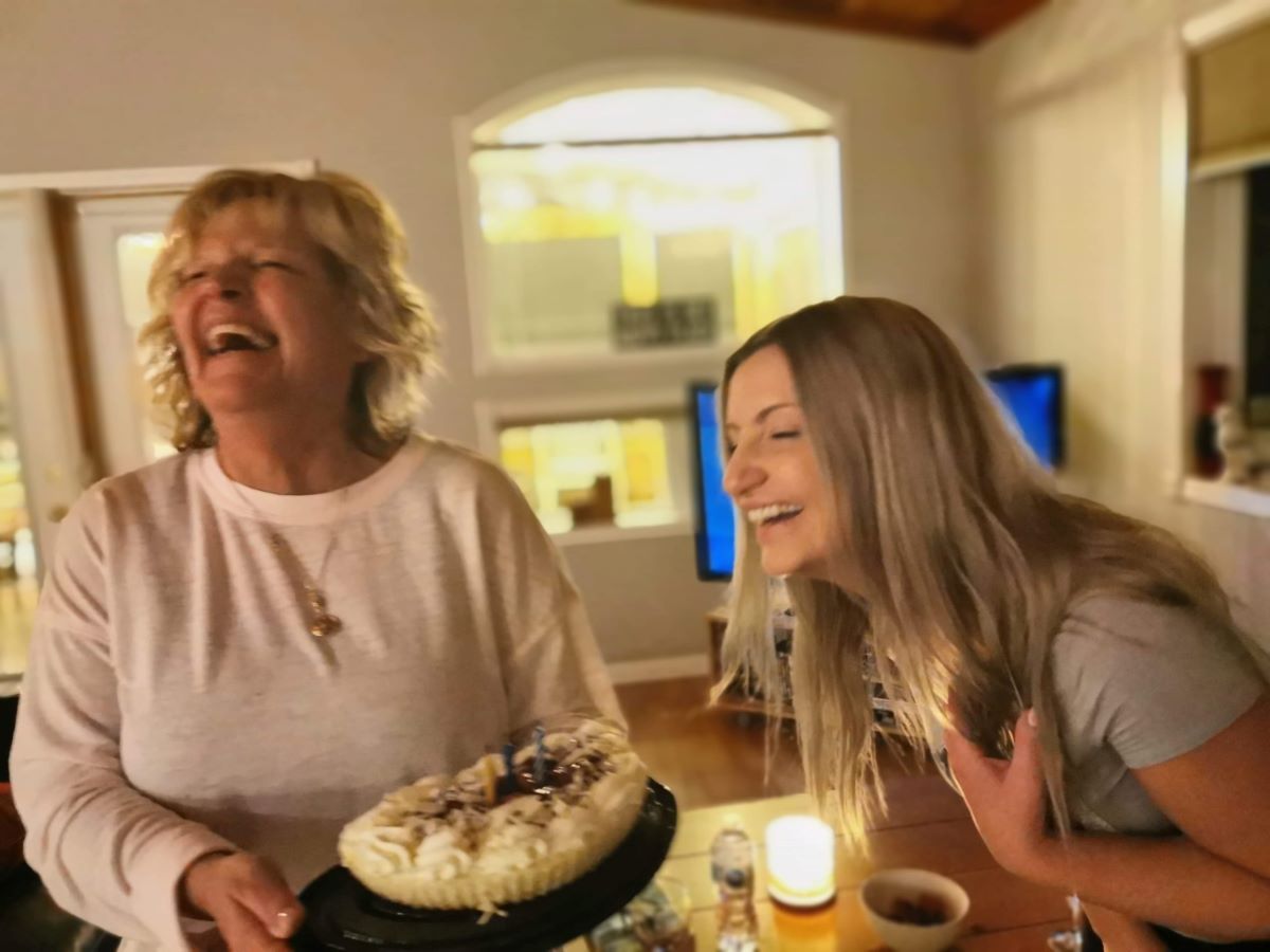 An older woman with short curly blond hair and a light skin tone is slightly out of focus. She’s laughing and holding a cake with unlit candles. To her left, a younger blond woman with a light skin tone, also laughing, leans in towards her mother and the cake.