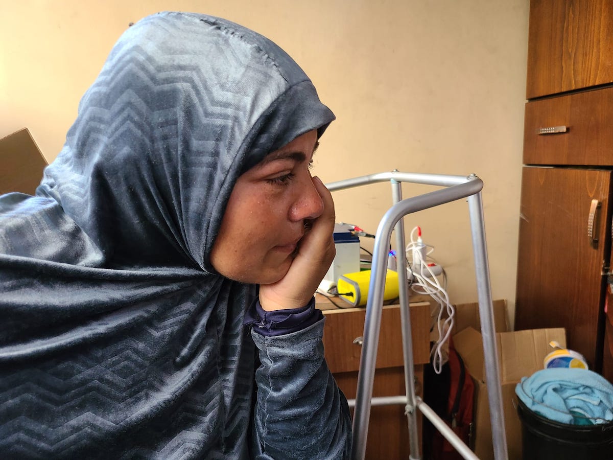 A woman with a blue head covering looks towards the right of the frame, her head resting on her hand. She has tears in her eyes.