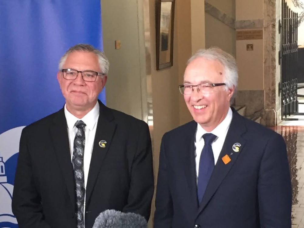 Two older men with light-toned skin and grey hair, wearing dark suits, white shirts and ties stand smiling.