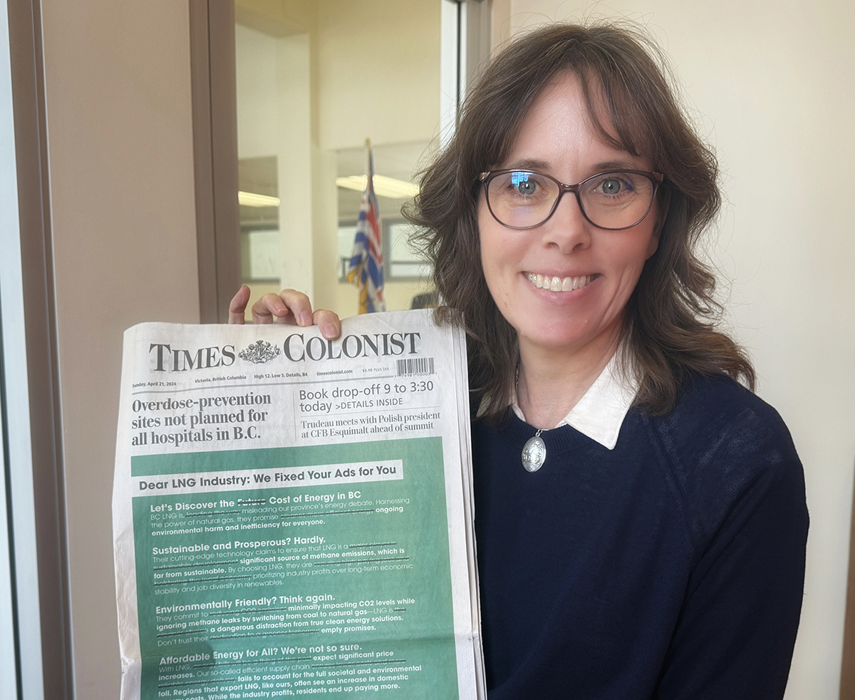 A woman with dark hair and classes smiles at the camera as she holds a newspaper with a large green advertisement on the front page.