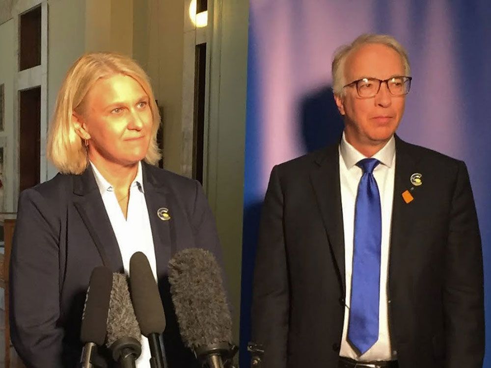 A light-skinned blonde woman wearing a blue suit stands beside an older light-skinned man with grey hair and glasses, wearing a dark suit, white shirt and blue tie.