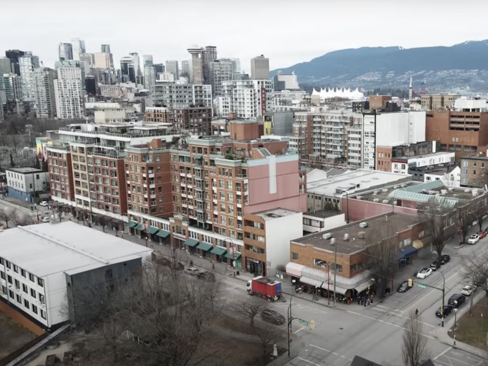 An aerial shot of Union Street and Gore Avenue in Vancouver&rsquo;s Chinatown features a large multi-storey brown brick apartment building with green storefront awnings and other small businesses with colourful awnings on the street. Cars are parked on the sides of the quiet streets and the sky is grey.