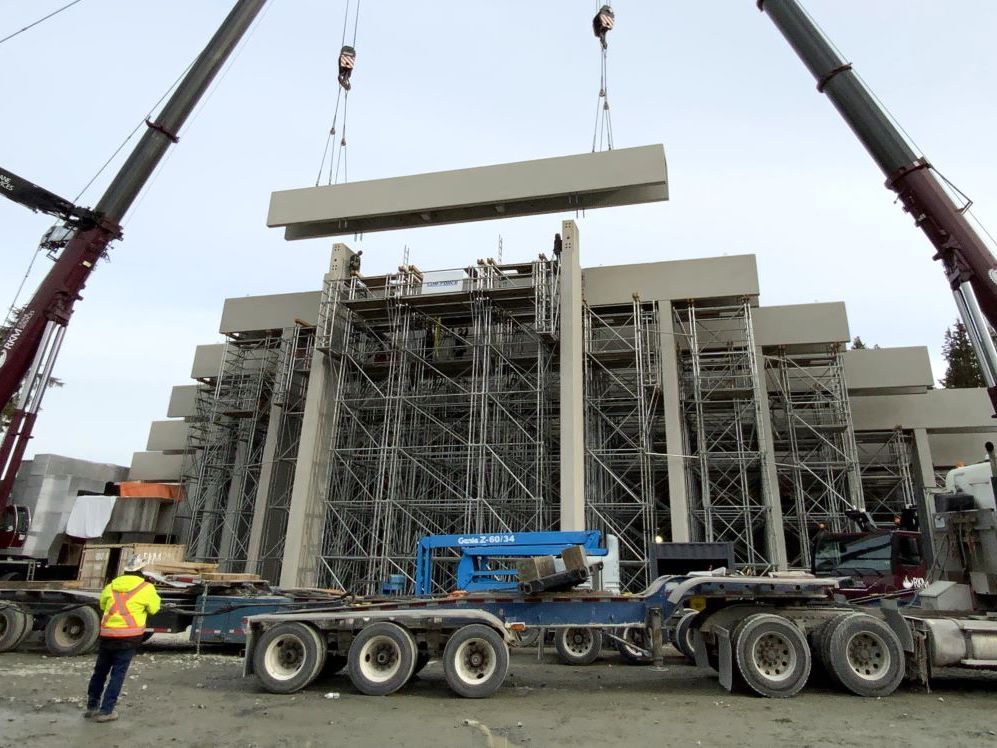 A beam is being lowered onto a scaffolding-supported structure under construction. Two crane arms can be seen on either side of the frame.