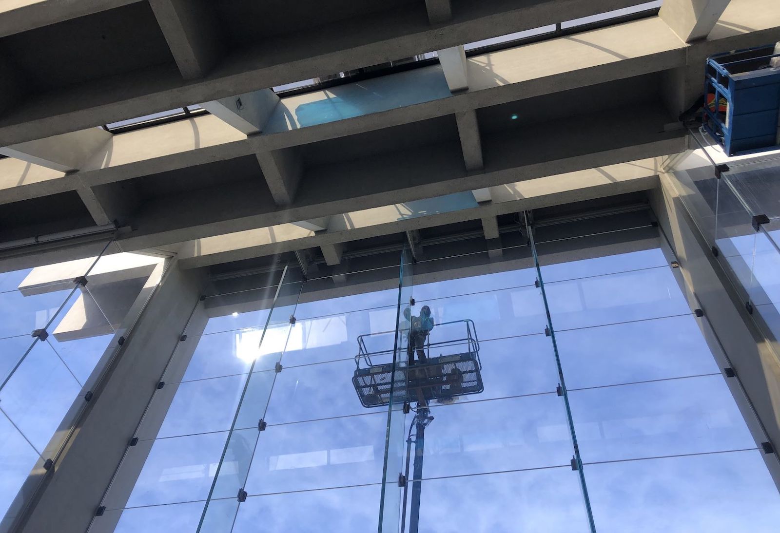 A tall wall of clear glass panes framed by bare concrete columns and a concrete beam roof. A person stands high on a movable platform outside the glass.