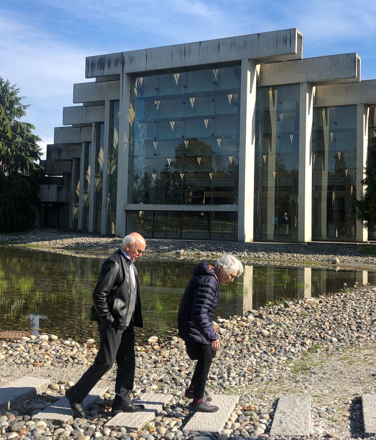 An elderly man and woman walk on a flagstone and stone path in front of a building made of glass panes and concrete beams.
