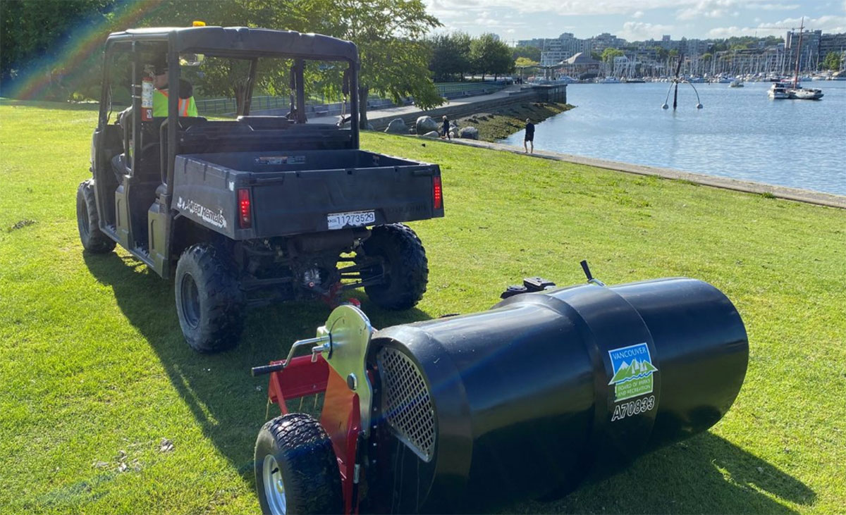 A parks vehicle tows a cylindrical black contraption with a Vancouver Board of Parks and Recreation logo along the grass by the water.