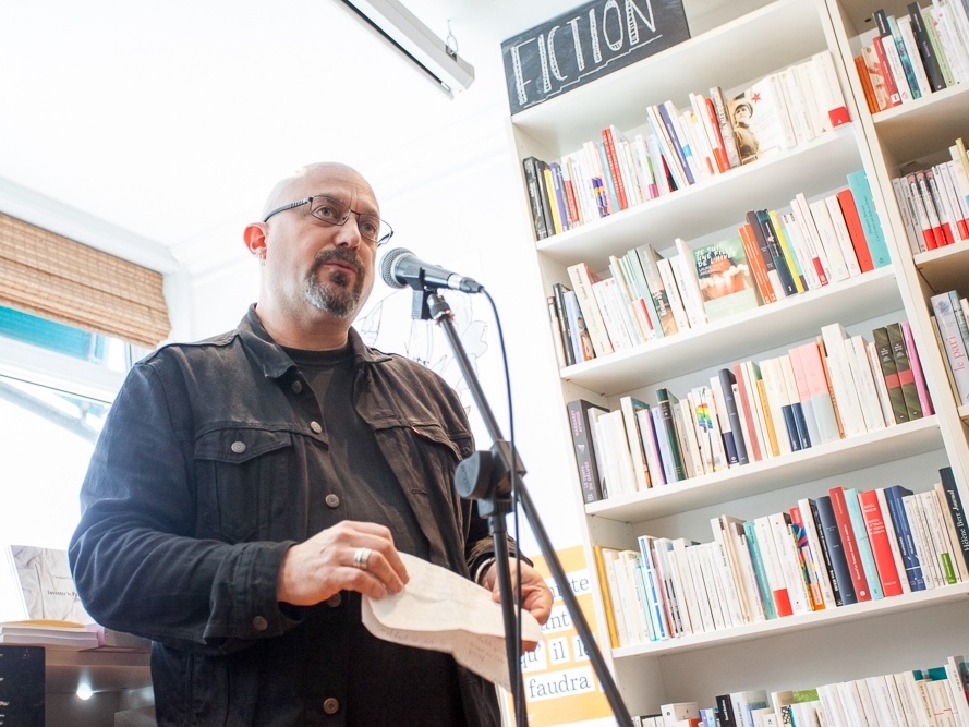 A bald middle-aged man with light skin, a beard and glasses wears a black jean jacket over a dark T-shirt. He stands at a microphone in a bookstore. Behind him, bright daylight is streaming through the windows.